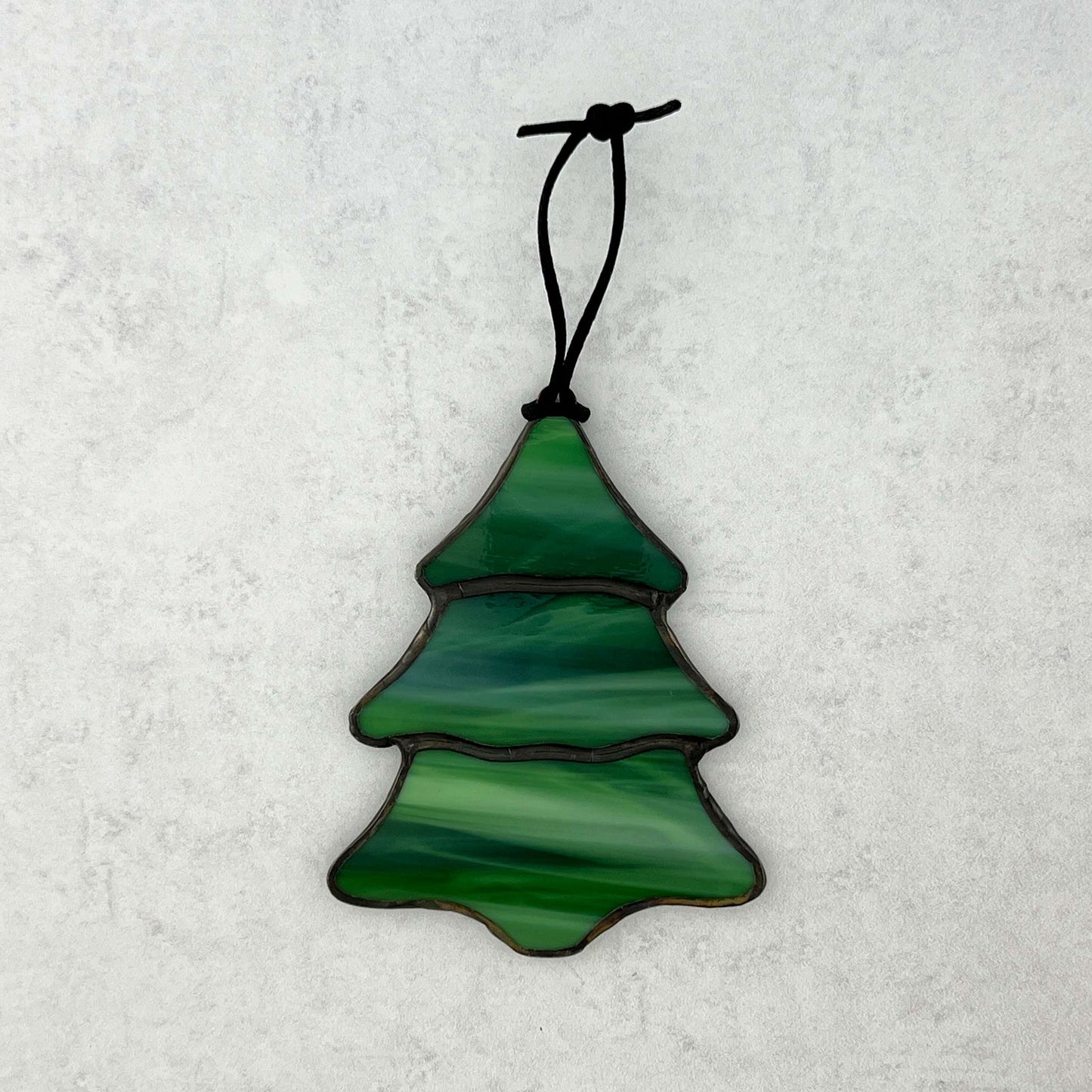 A stained glass multi-colored green Christmas tree ornament against a white background. There are swirls of white in the green glass that make it feel like snow is falling on the tree. A black cord is shown for hanging