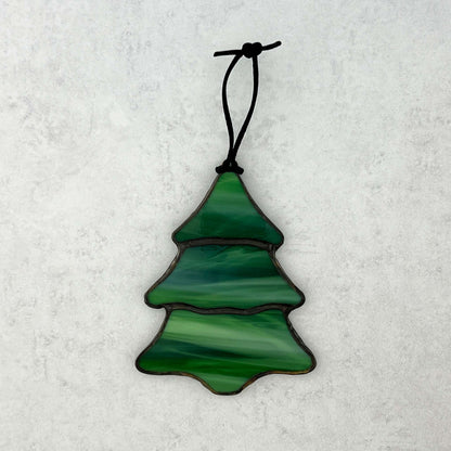 A stained glass multi-colored green Christmas tree ornament against a white background. There are swirls of white in the green glass that make it feel like snow is falling on the tree. A black cord is shown for hanging