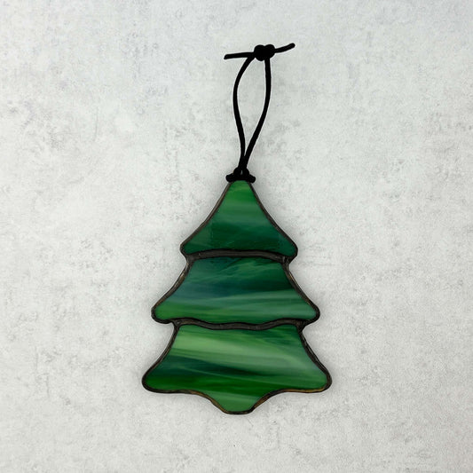 A stained glass multi-colored green Christmas tree ornament against a white background. There are swirls of white in the green glass that make it feel like snow is falling on the tree. A black cord is shown for hanging