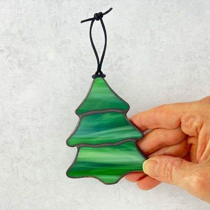 Hand holding a stained glass multi-colored green Christmas tree ornament against a white background. There are swirls of white in the green glass that make it feel like snow is falling on the tree.