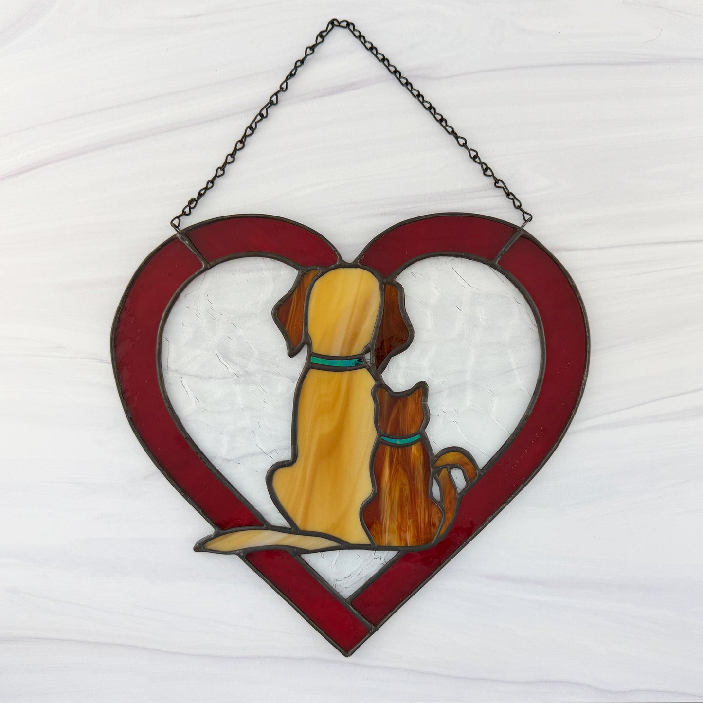 Stained glass heart-shaped window hanging with a dog and cat design on a white background. The dog is amber and the cat a ginger color.