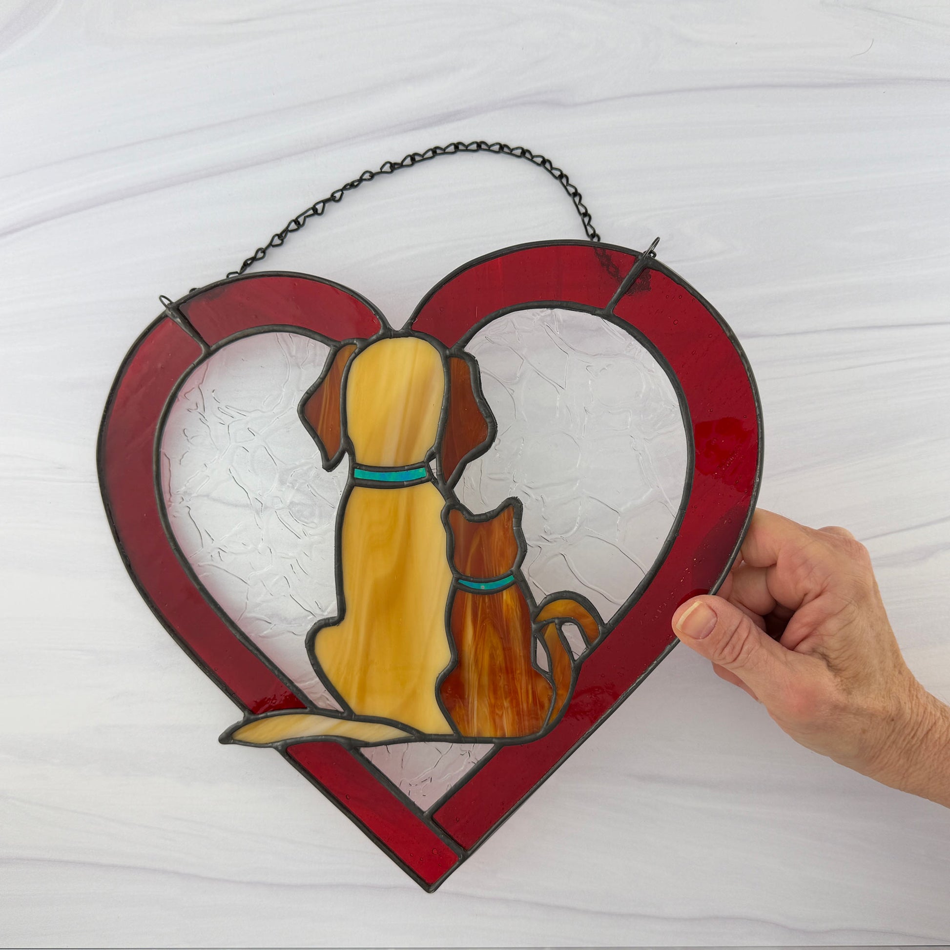 Stained glass heart-shaped panel with a dog and cat sitting together, held by a hand on a white background. Both pets have matching blue collars.