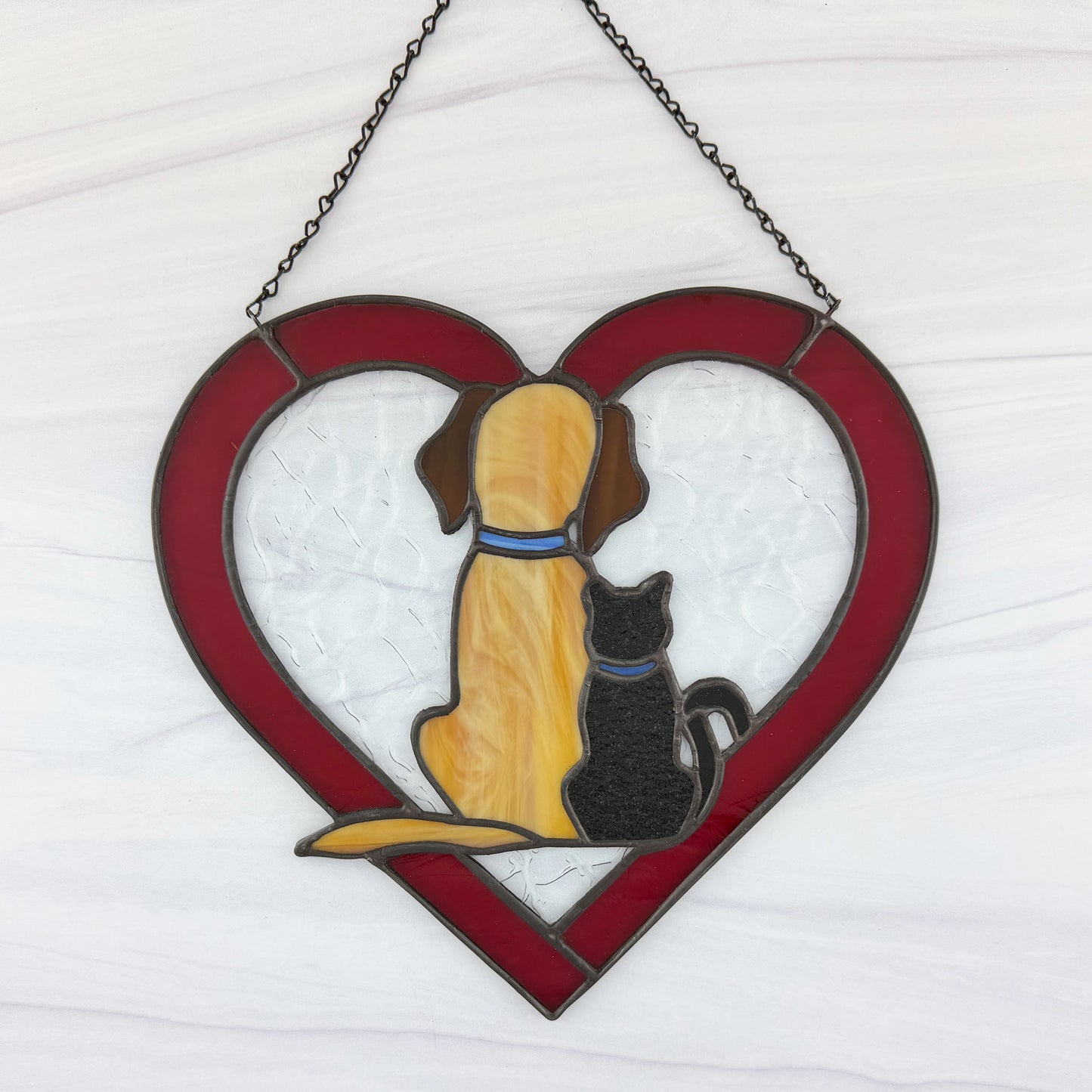 Stained glass red heart with a dog and cat design on a white background. A beige colored dog with brown ears is sitting next to a black cat. Both have blue collars.