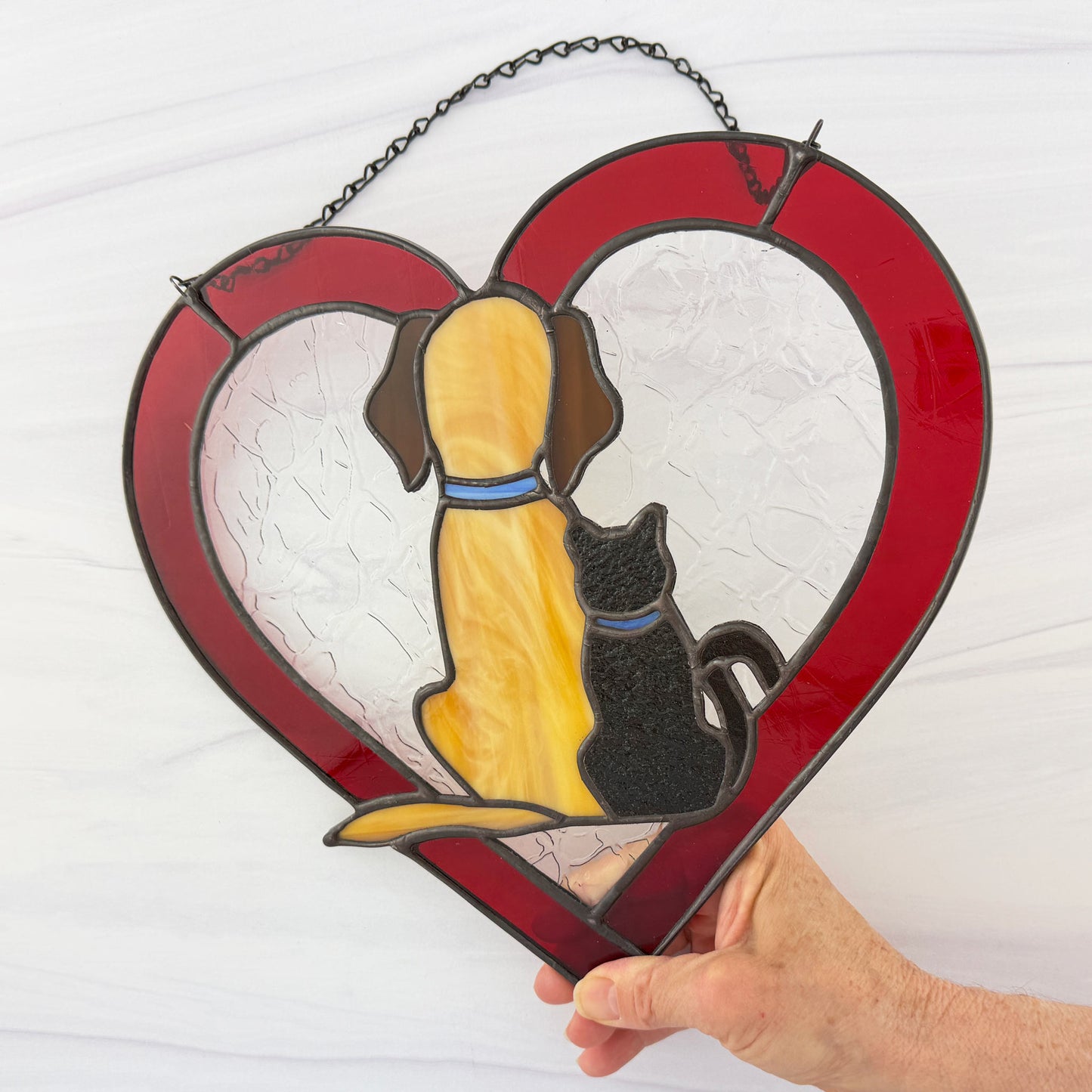 Red Heart-shaped stained glass panel with a dog and cat design, held by a hand against a white background.