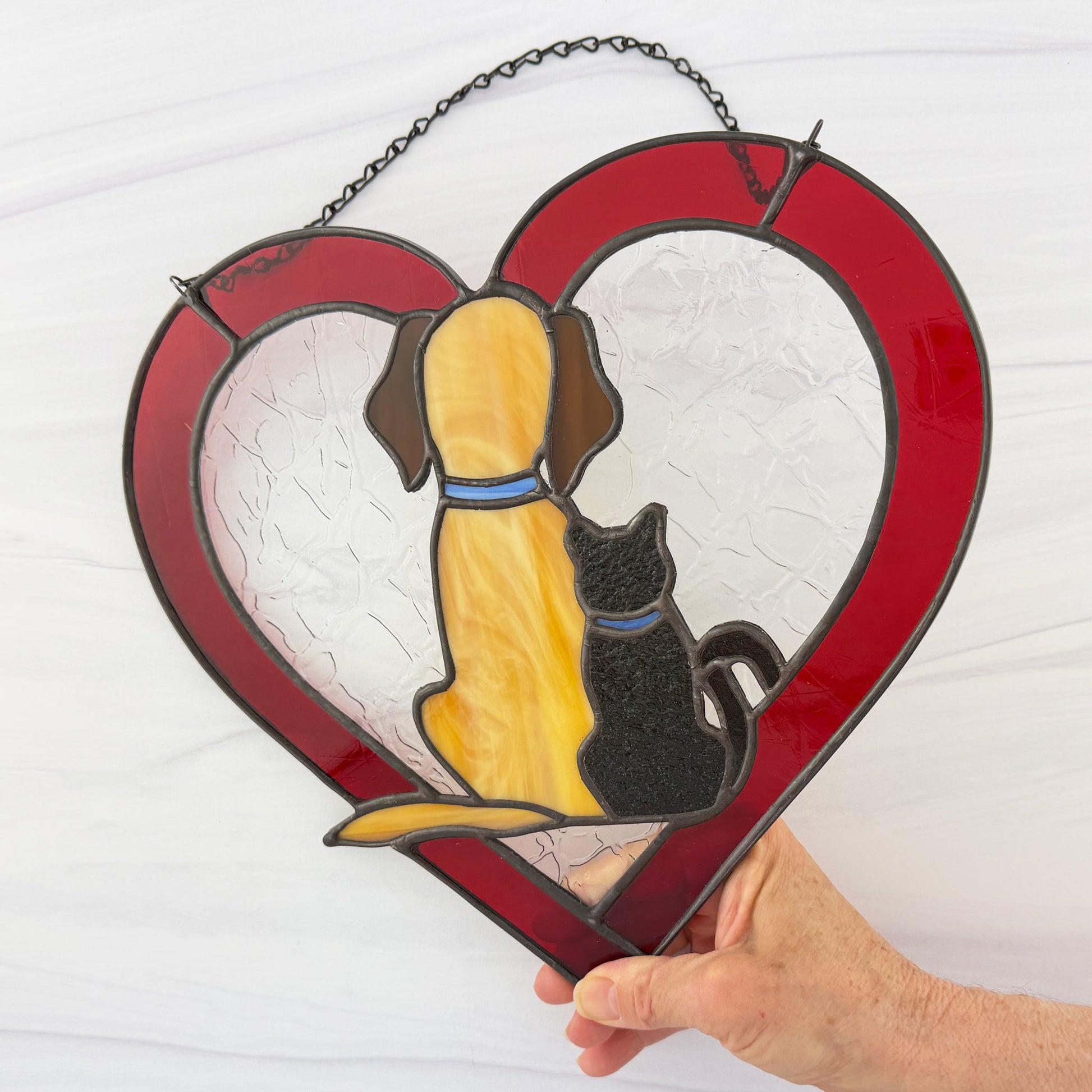 Red Heart-shaped stained glass panel with a dog and cat design, held by a hand against a white background.