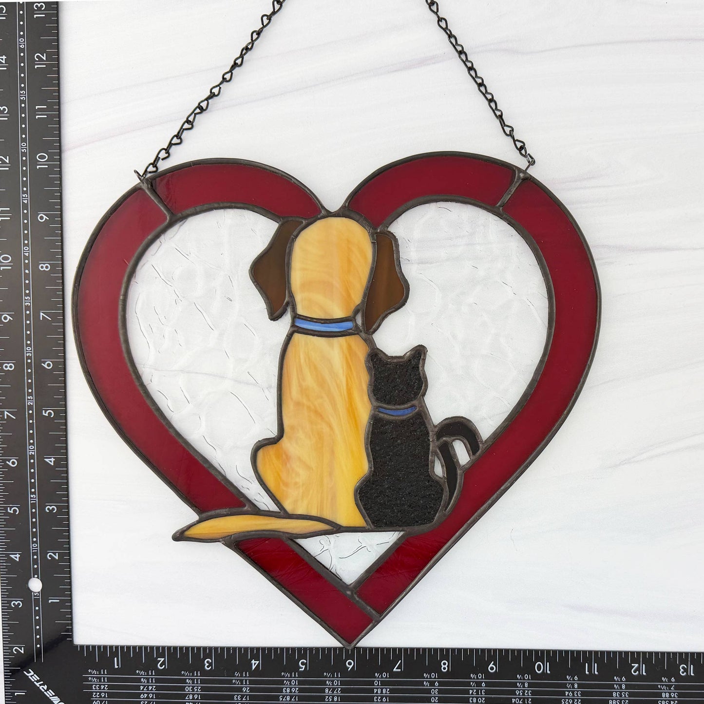 Stained glass suncatcher with a dog and cat in a red heart shape, hanging on a white background with a ruler for scale.