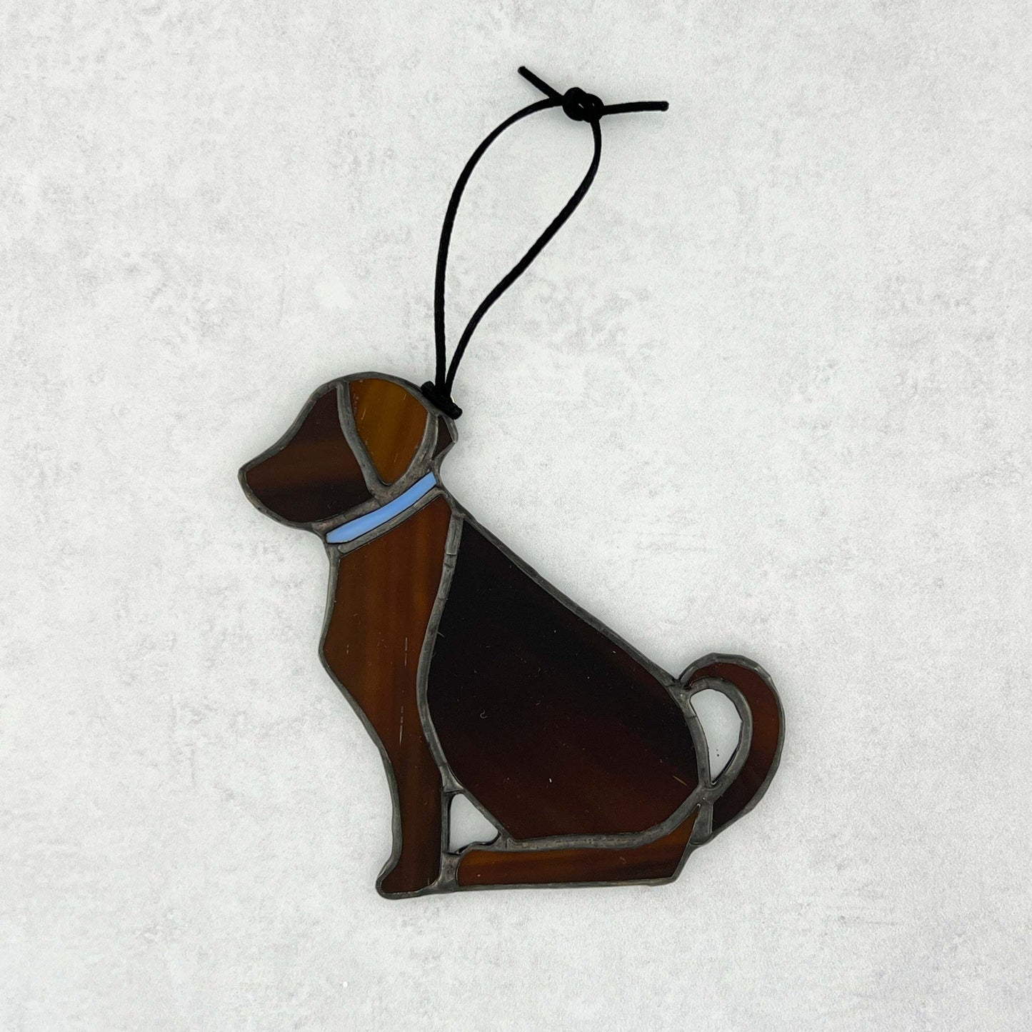 Decorative brown stained glass dog ornament with a blue collar on a snowy background.  The dog is sitting with his tail curled up behind him. A black cord is used for hanging.  The ear and legs are a lighter brown than the body.