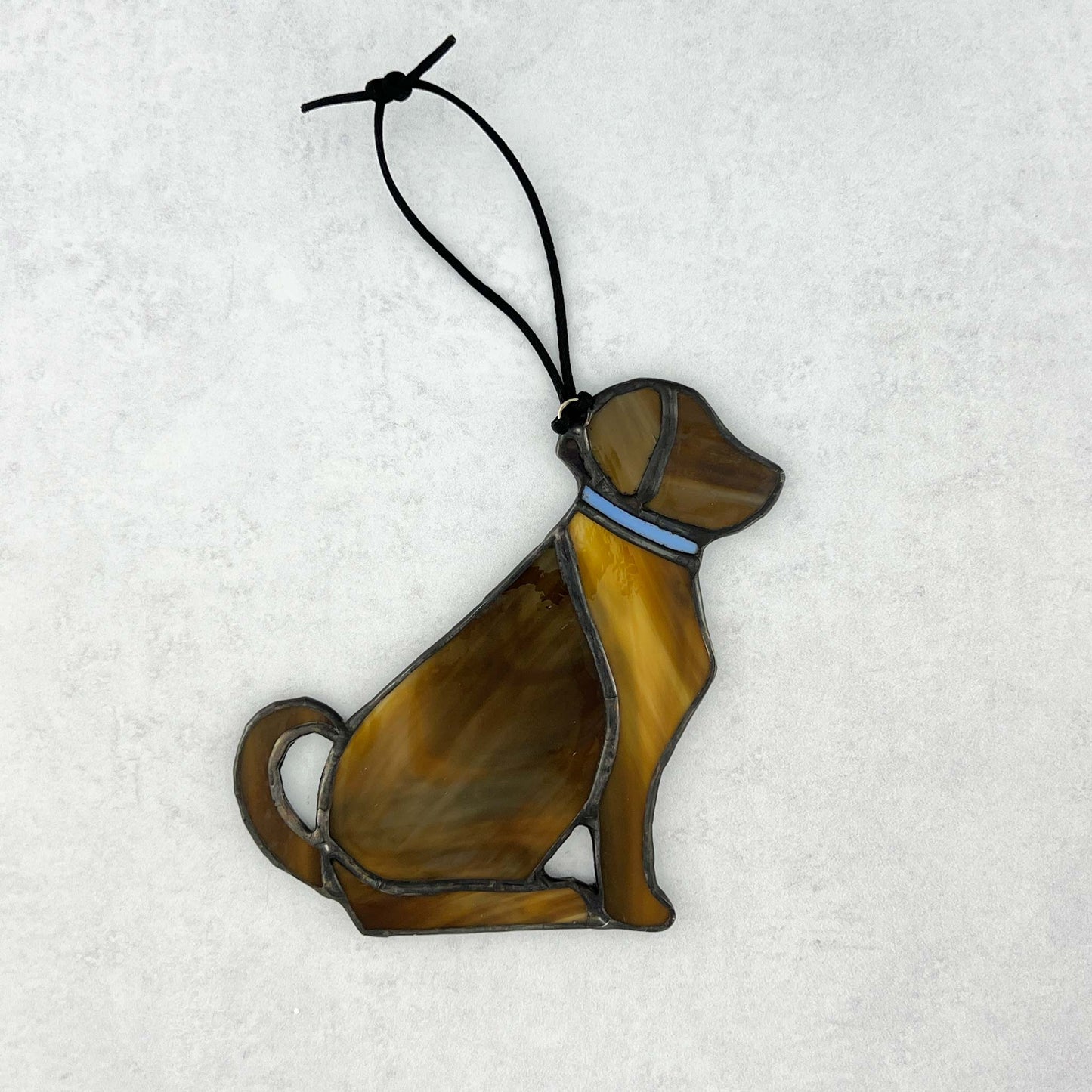 The back side of a decorative brown stained glass dog ornament with a blue collar on a snowy background.  The dog is sitting with his tail curled up behind him. A black cord is used for hanging.  