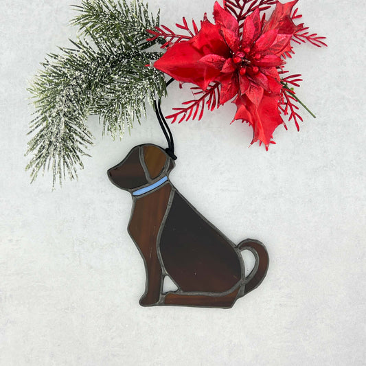 Decorative brown stained glass dog ornament with a blue collar on a snowy background.  The dog is sitting with his tail curled up behind him. A black cord is used for hanging.  