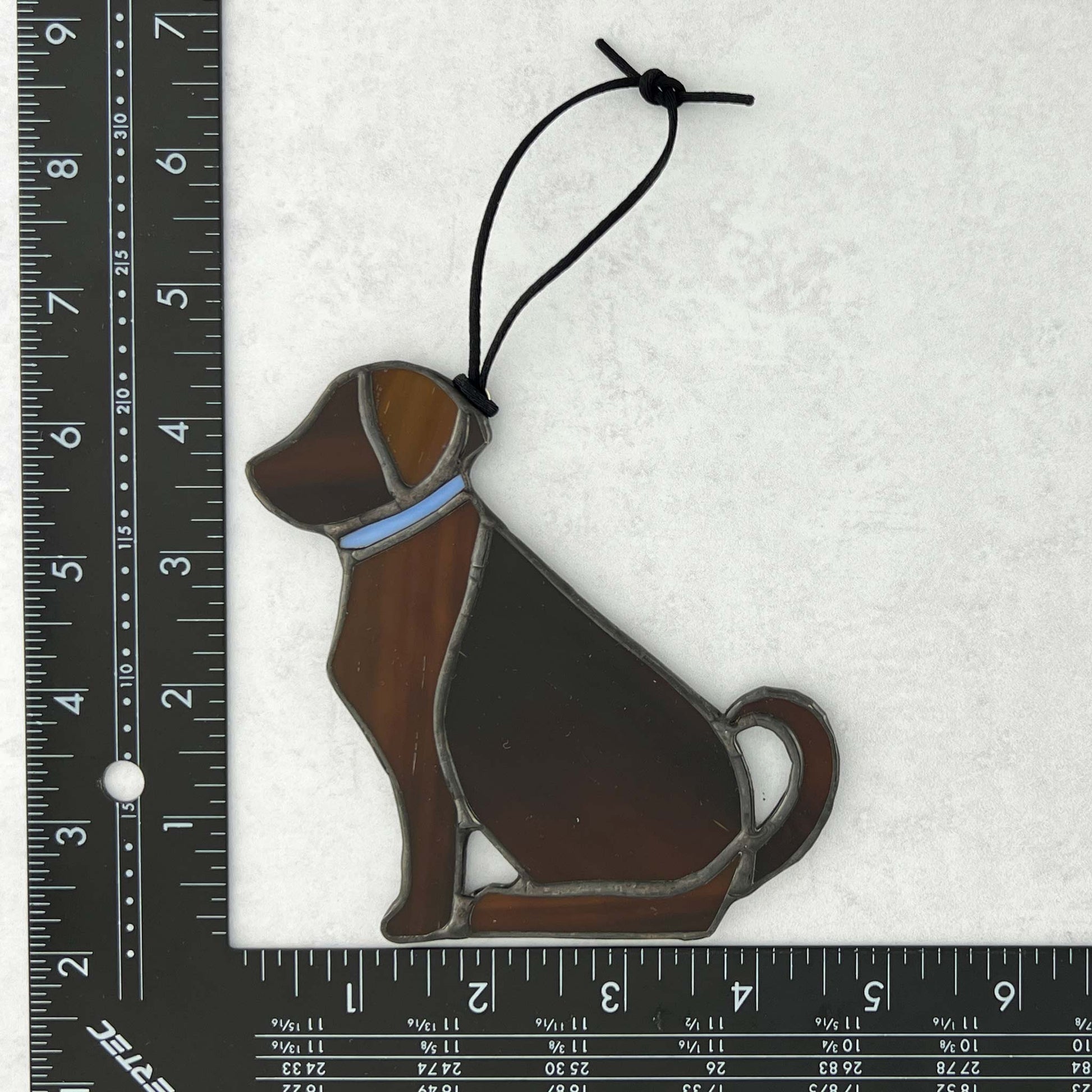 Stained glass dog ornament with a blue collar on a white background with a ruler for scale.