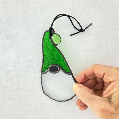 Hand holding a glass ornament gnome with a green hat on a light gray background