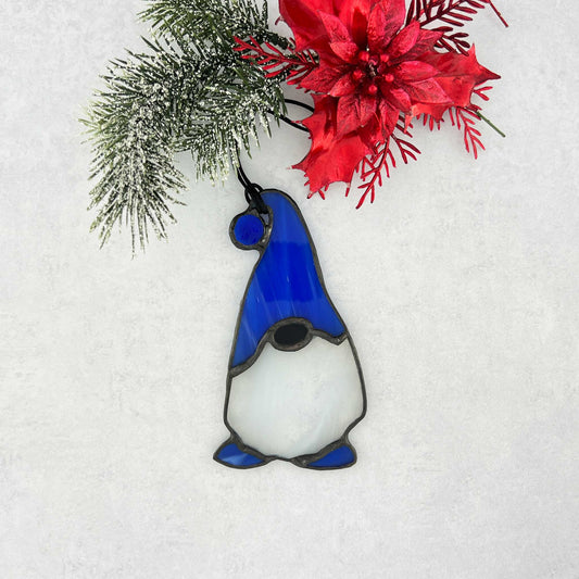 Stained glass gnome with a blue hat featuring a blue ball at the end and complimented by blue feet.  He has a beard made of white and clear glass. Shown hear hanging from a pine tree.