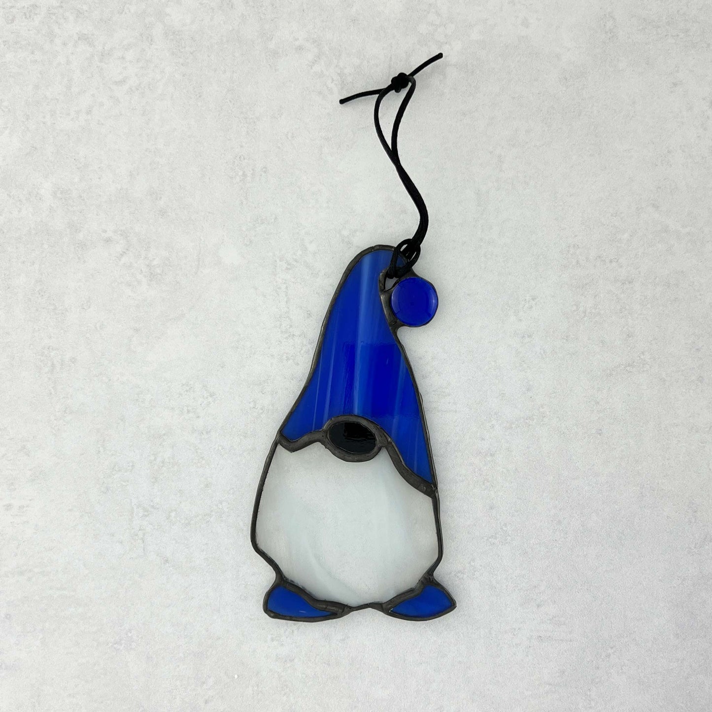 Stained glass gnome with a blue hat featuring a blue ball at the end and complimented by blue feet.  He has a beard made of white and clear glass.
