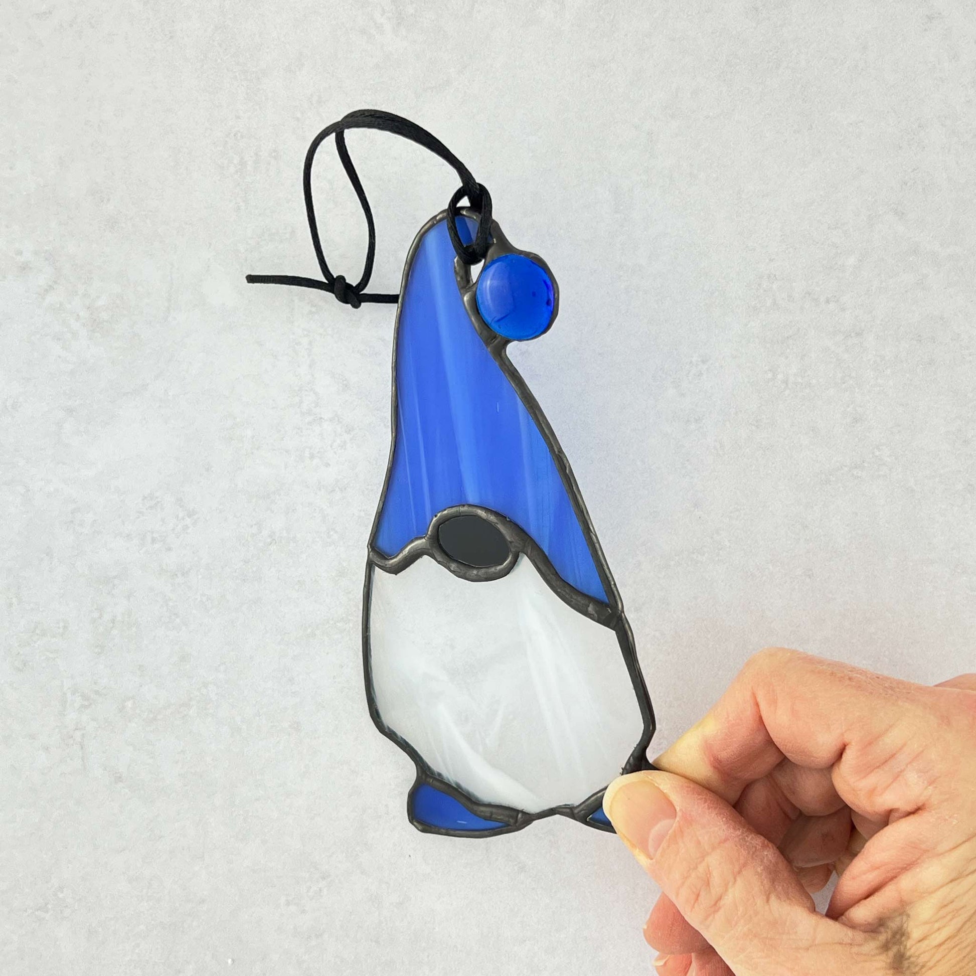 Hand holding a Stained glass gnome with a blue hat featuring a blue ball at the end and complimented by blue feet.  He has a beard made of white and clear glass.