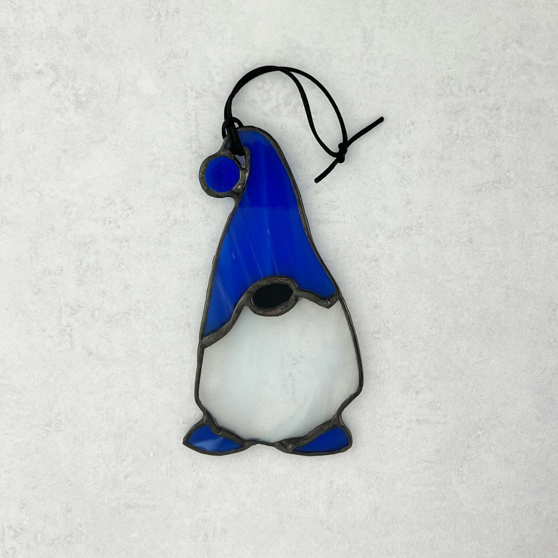 the back side of a Stained glass gnome with a blue hat featuring a blue ball at the end and complimented by blue feet.  He has a beard made of white and clear glass.