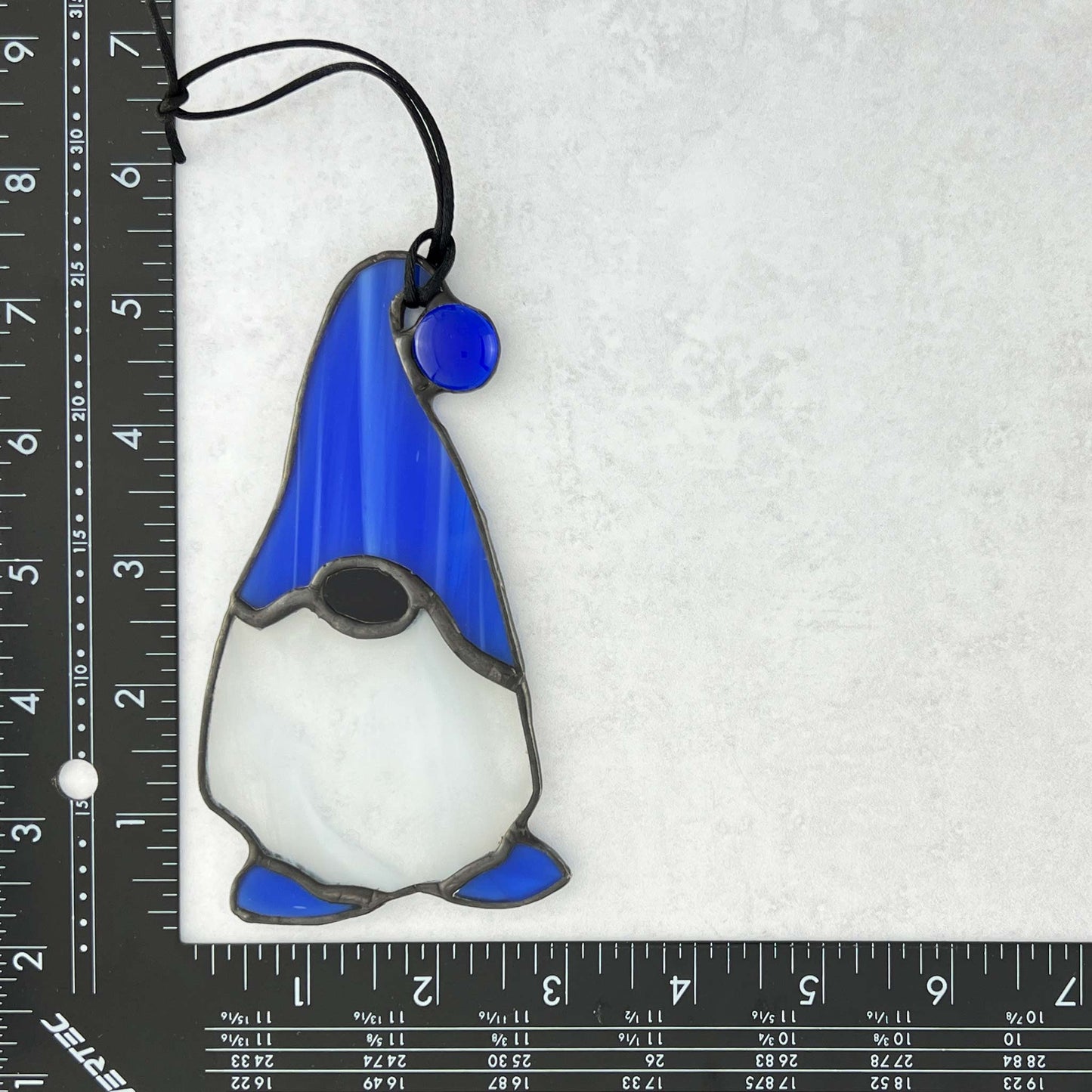 Stained glass gnome with blue hat and white face on a white background with ruler for scale.