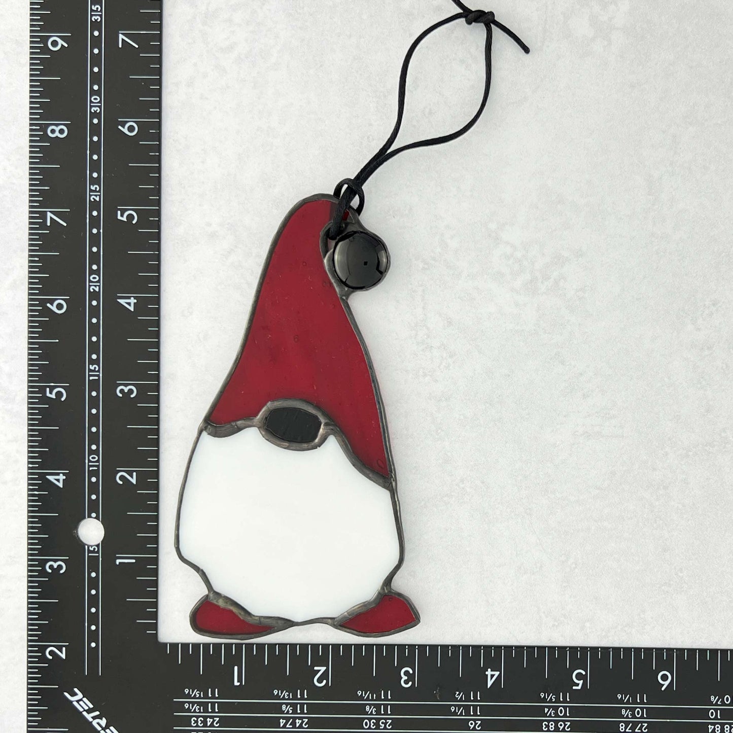 Stained glass gnome ornament with red hat and shoes on a white background with ruler for scale.