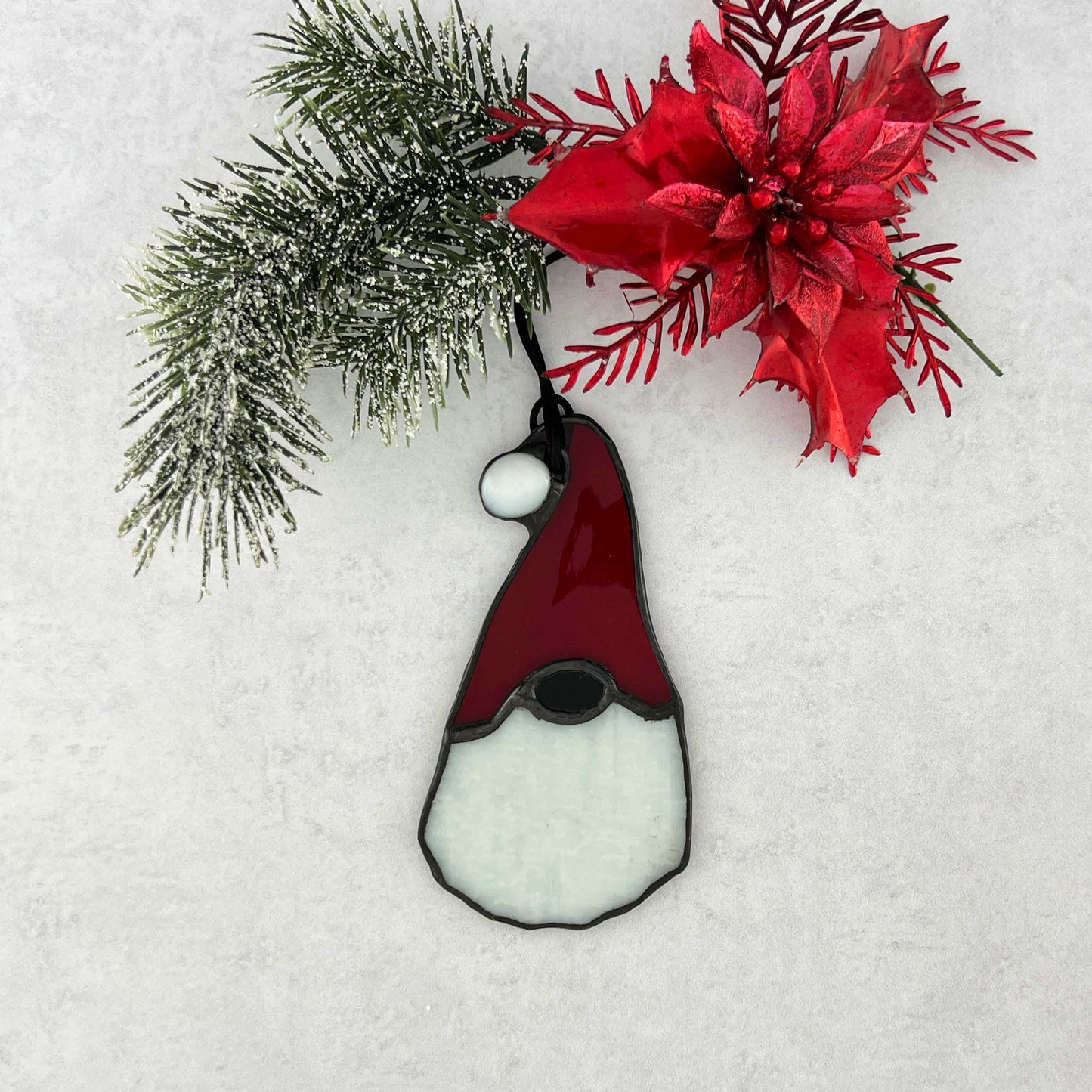 Stained glass gnome ornament with red hat that has a raised white ball on the end and white textured beard on a snowy background with poinsettia and pine branch.