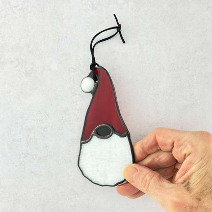 Hand holding a Stained glass gnome ornament with red hat and white beard on a textured white background. The hat has a white ball at the end.
