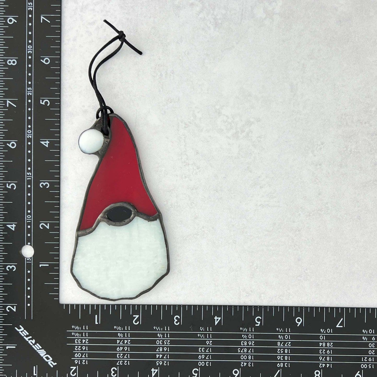 Stained glass gnome ornament with red hat and white body with ruler for scale.
