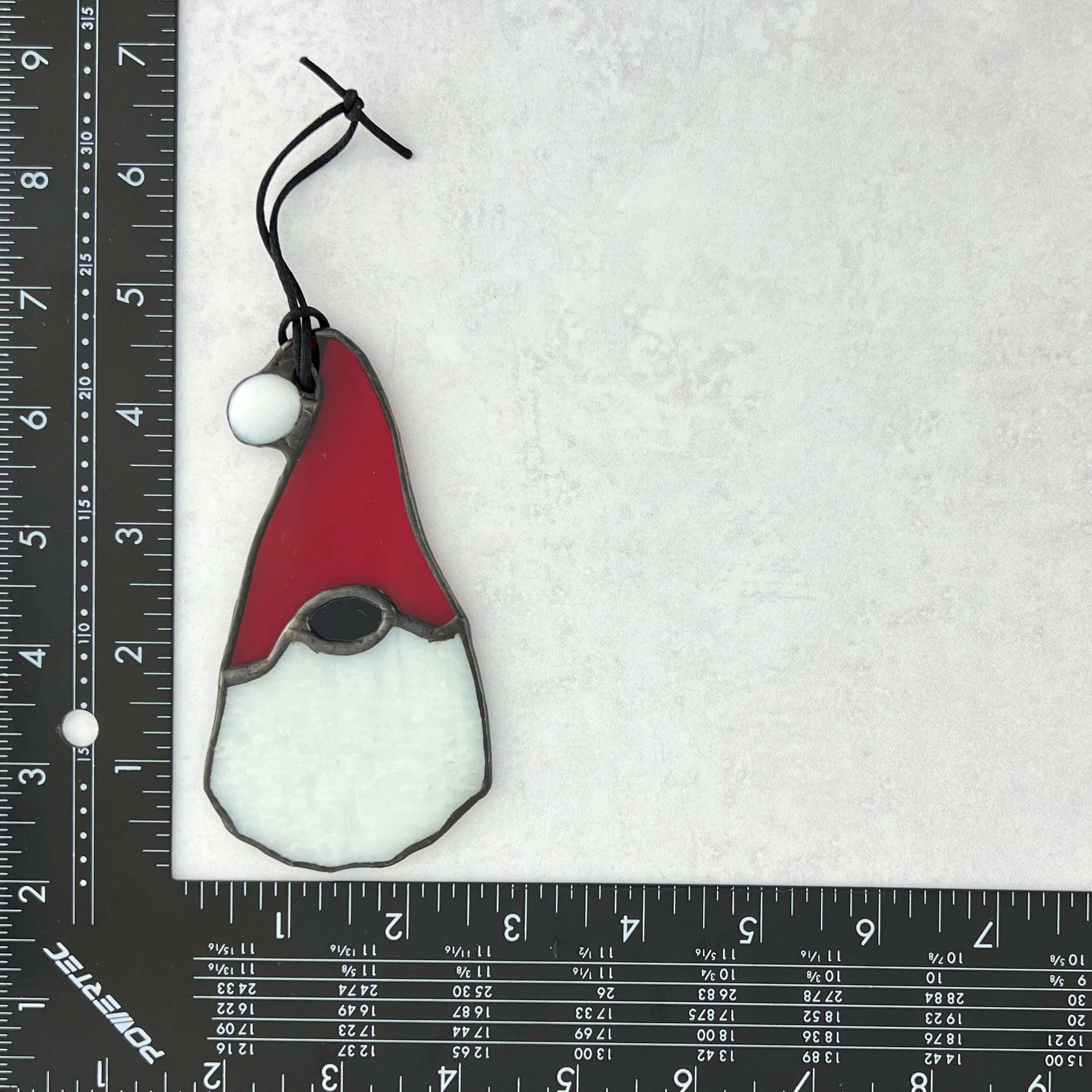 Stained glass gnome ornament with red hat and white body with ruler for scale.