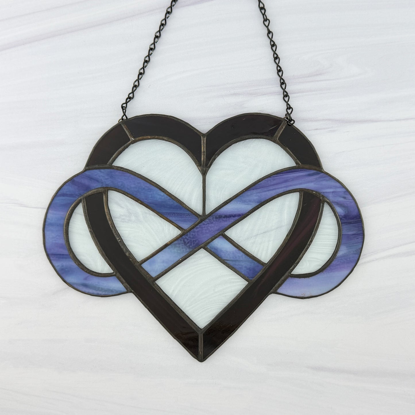 Stained glass dark purple heart with light puple-blue infinity symbol on a white background. Clear wavy glass is between the open areas and it is hung from a black chain.