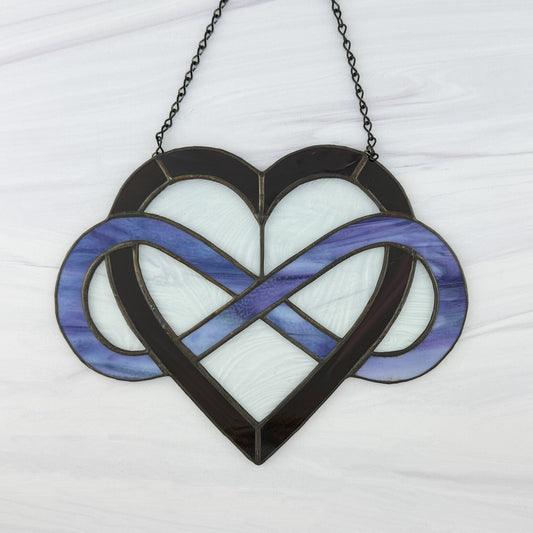 Stained glass dark purple heart with light puple-blue infinity symbol on a white background. Clear wavy glass is between the open areas and it is hung from a black chain.