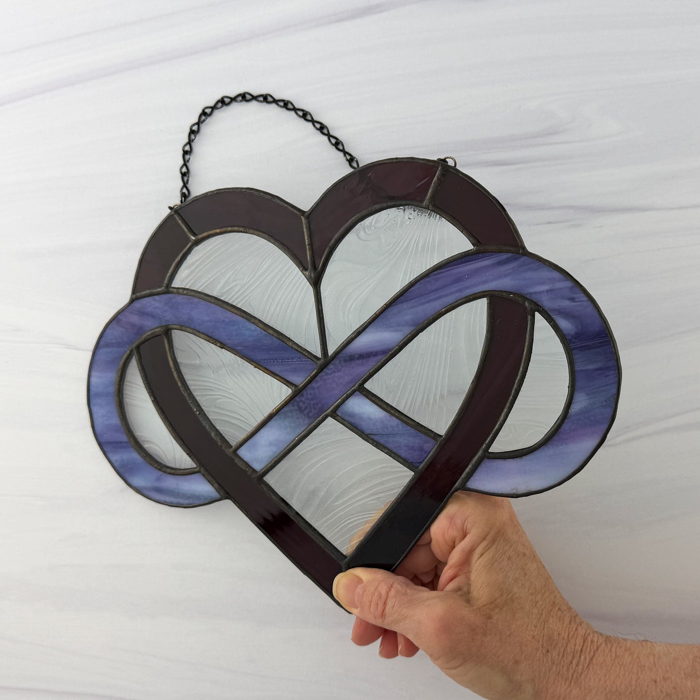 Dark purple Heart stained glass panel with intertwined infinity design held by a hand against a white background