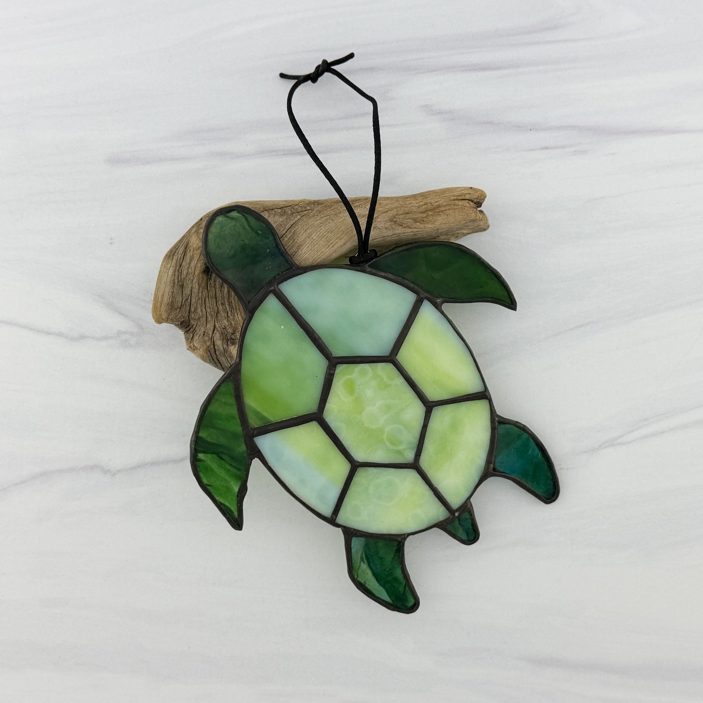 Stained glass sea turtle suncatcher resting on driftwood on a white background. The shell is a mottled light green opaque glass and the flippers and head are a translucent blue-green glass
