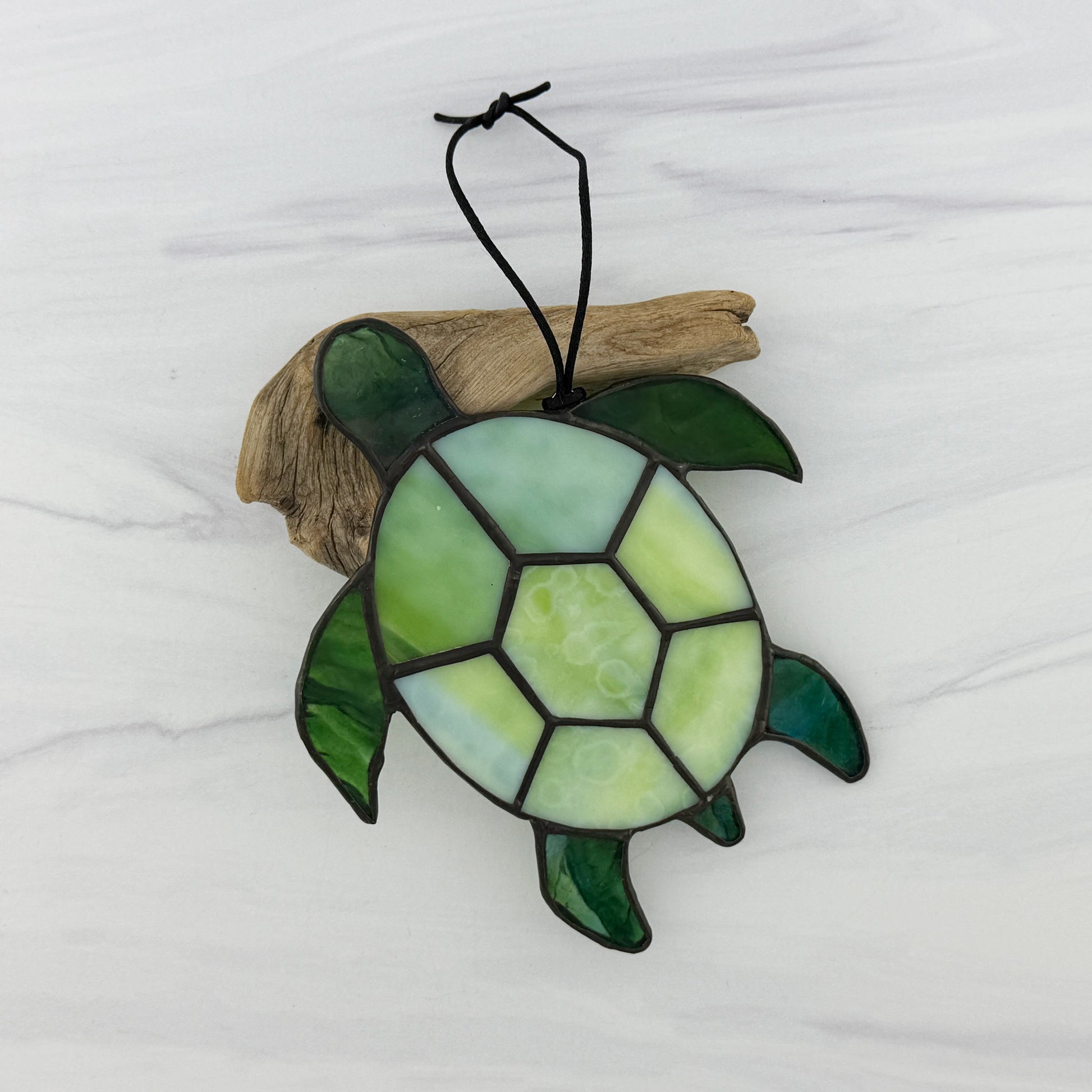 Stained glass sea turtle suncatcher resting on driftwood on a white background. The shell is a mottled light green opaque glass and the flippers and head are a translucent blue-green glass