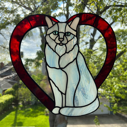 An all white stained glass sitting cat surrounded by a red heart. The glass used for the white fur is textured to resemble real fur and the eyes are an iridescent blue.  Light pink is used for the inner ear and the nose. A clear rainy glass is used between the cat and the heart for stability.