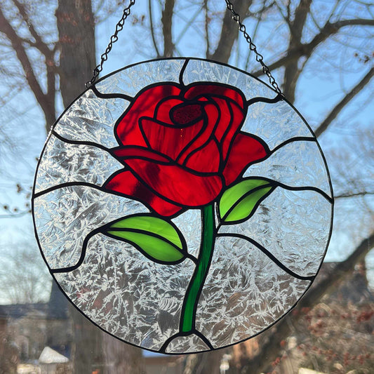 This red rose stained glass suncatcher is a stunning piece, perfect for brightening up any room. Measuring 8.5 inches, this suncatcher features a vibrant red rose at its center. The rose is a mix of red shades, and the leaves are light green. Surrounding the rose is a delicate clear “fern-like” glass, adding elegance.